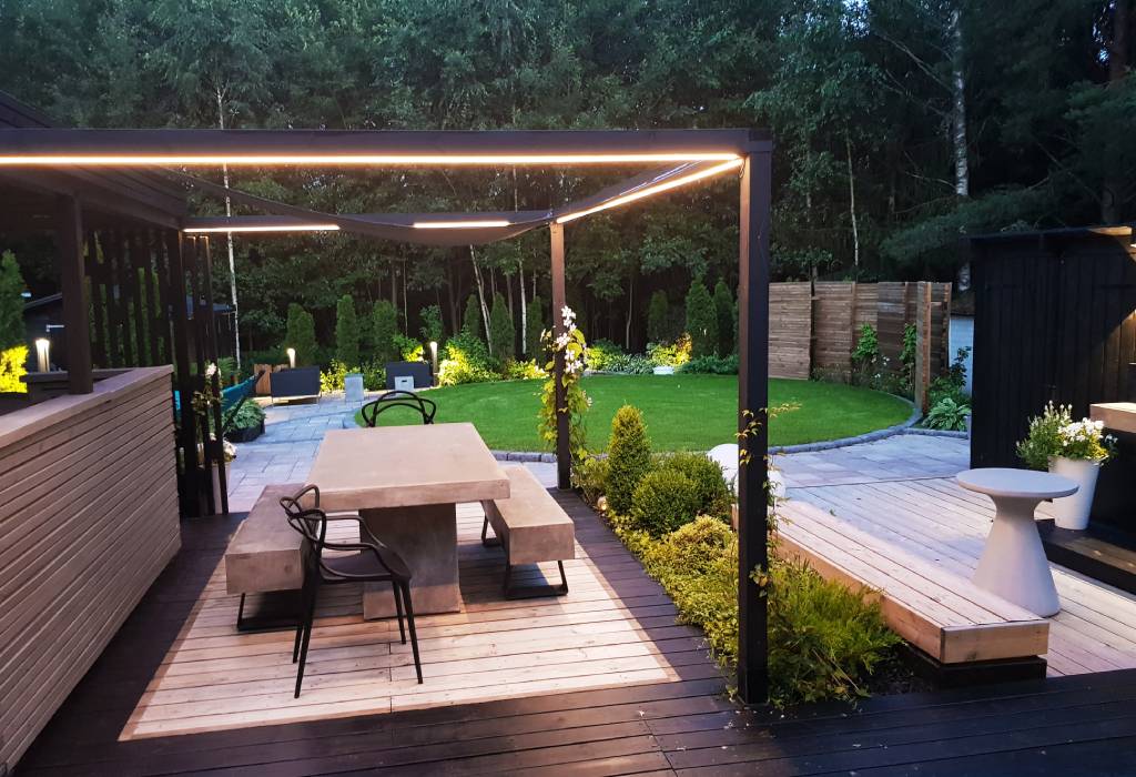 Moderni ulkoruokailutila, jossa on pergola varustettuna pehmeällä LED-valaistuksella, betonipöytä penkkeineen ja tuoleineen. Alue on vehreän nurmikon ympäröimä ja taustalla kohoavat komeat puut. Puuterassilla on käytännöllinen oleskelualue sekä pieni puutarha täydentämässä tunnelmaa. Tutustu upeisiin LED-valaisimiin LedStore.fi-verkkokaupassamme ja luo oma unelmiesi ulkotila!