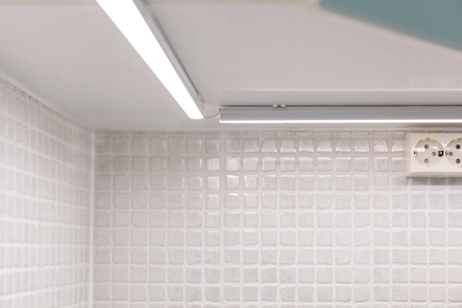Close-up of tiled wall with LED fluorescent lights under a cabinet, illuminating the space. Visible electrical outlet. Neutral, minimalist design.